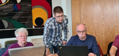 Zwei Teilnehmer des Kurses sitzen vor ihren Laptops, in der Mitte steht Herr Decker und erklärt einem Teilnehmer etwas am Computer.