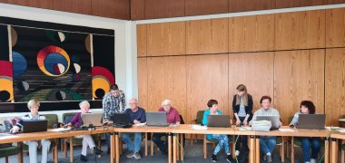 Der große Ratssaal mit einer langen Tischreihe an denen die Teilnehmer*innen des Einsteigerkurses mit ihren Laptops sitzen. Zwischendrin stehen Herr Decker und Frau Marotte und helfen den Teilnehmer*innen bei der Bedienung der Computer.
