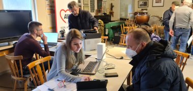 Raum der Handyhilfe, dort stehen mehrere Tische und Stühle. An einem Tisch sitzt Frau Marotte mit einem Mann. Frau Marotte versucht die Probleme vom Tablet des Mannes zu lösen und erklärt dabei ihr Vorgehen. Herr Decker sitzt an einem weiteren Tisch und hilft einem anderen Mann der Probleme mit seinem Drucker hat.