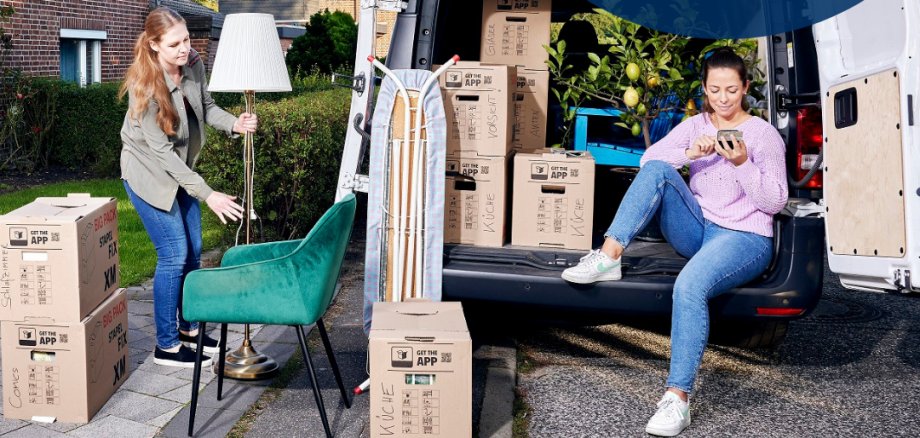 Neues Zuhause? Online anmelden. Digitale Ummeldung ab sofort auch für Familien! wohnsitzanmeldung.de Zwei Frauen am Tag am Bürgersteig in einer Wohngegend. Um sie herum  ein Lieferwagen und Umzugskartons. Weitere Haushaltsgegenstände wie Sessel, Bügelbrett und Lampe stehen vor dem vollen Umzugswagen. Eine der Frauen sitzt auf der Pritsche des Wagens und tippt entspannt auf ihrem Smartphone. Die zweite Frau bringt eine Lampe.