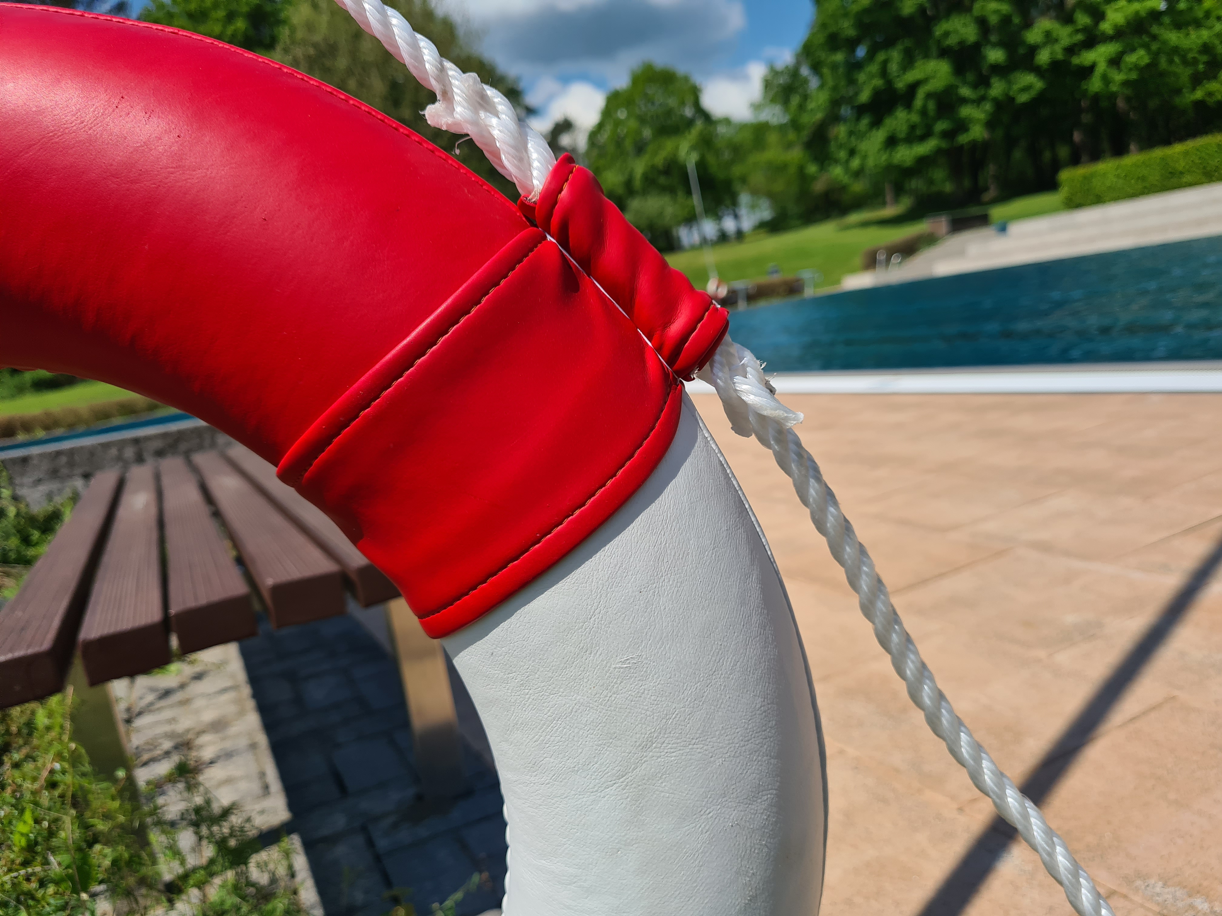 Rettungsring Freibad Im Vordergrund ist ein viertel eines Rettungsring und eine Holzbank zu sehen. Dahinter befindet sich ein Schwimmbecken und eine Liegewiese.