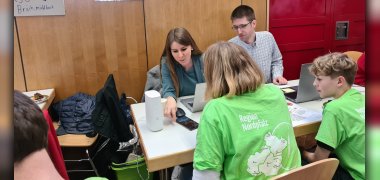 Frau Marotte und Herr Decker von der Handyhilfe sitzen an einem Tisch mit Laptops, gegenüber sitzen zwei Personen in grünen T-Shirts mit der Aufschrift "Region Nordpfalz".