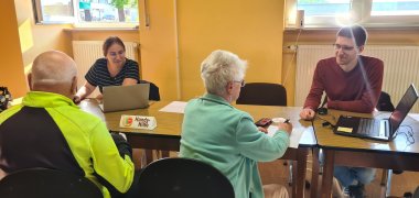 Hier ist ein Bild von der Handy Hilfe, es ist eine Tischreihe zusehen wo Frau Rimlinger einen Mann bei seinem Handy Problem hilft, und wie Herr Decker einer Frau etwas am Laptop zeigt. Außerdem ist auf dem Tisch ein kleiner Papieraufsteller mit der Aufschrift Handy Hilfe.
