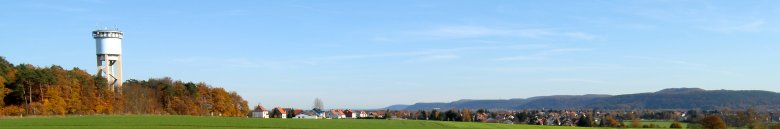 Lings ein Wasserturm im Wald. Davor ein Feld und daneben ein Dorf.