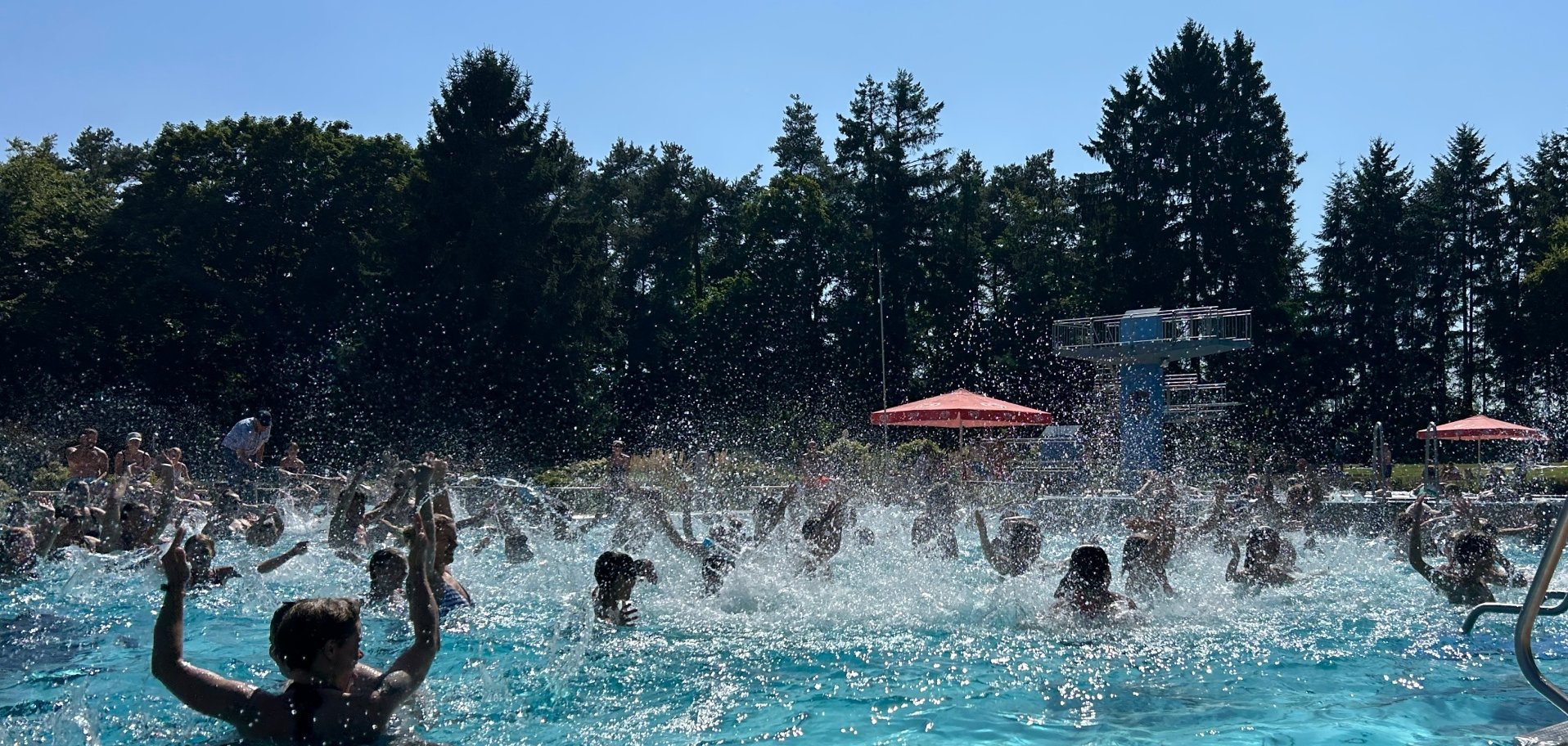 Ein turkisblaues Schwimmbad mit Badegästen in im Hintergrund grüne Bäume und blauer Himmel.